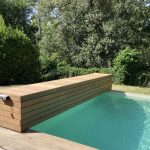 Volet hors sol de piscine enroulé et habillé d'un coffre en bois, situé dans un jardin avec la piscine ouverte en pleine journée.