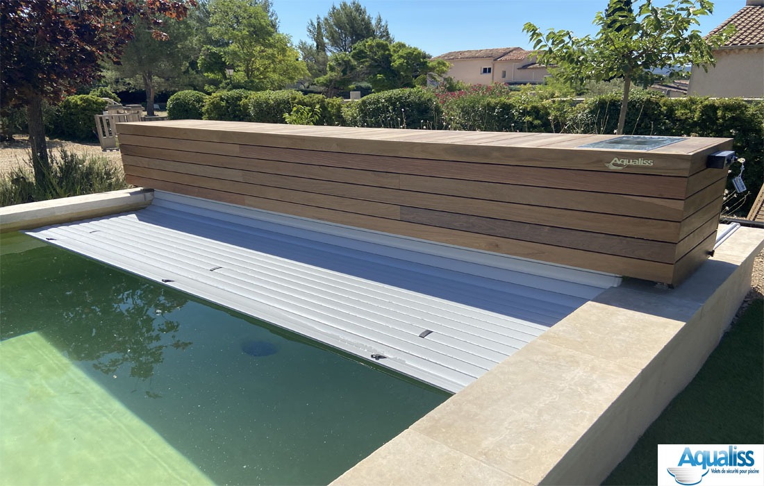 Couverture de piscine sur rails avec coffre en bois, tablier de lames se déployant dans le bassin, en plein jour.